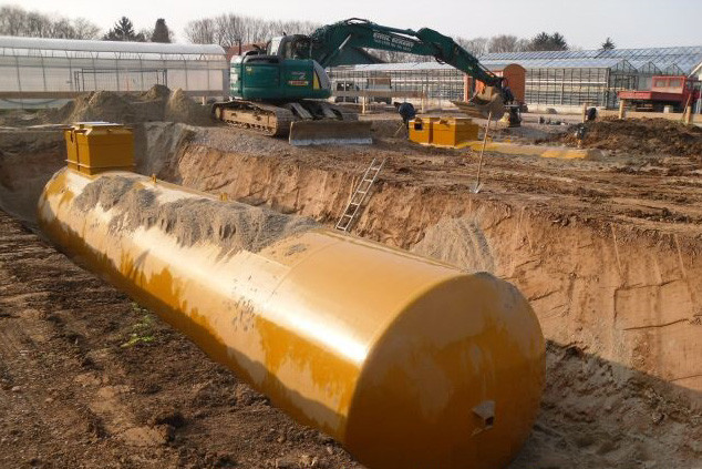 Erdtank in Baugrube mit Sand teilverfüllt, im Hintergrund Bagger auf Baustelle.