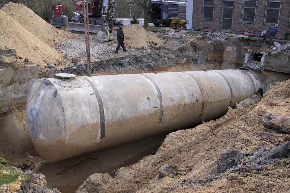 Alter Erdtank wird aus einer Baugrube gehoben, Arbeiter und Bagger im Hintergrund