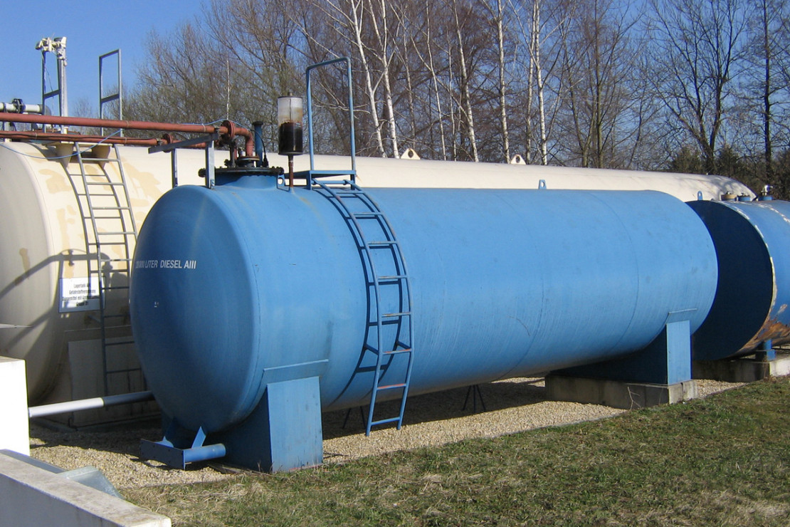blauer länglicher Tank, der auf eine Fläche vor einem weiteren größeren weißen Tank steht. Im Hintergrund sind Bäume und blauer Himmel zu sehen