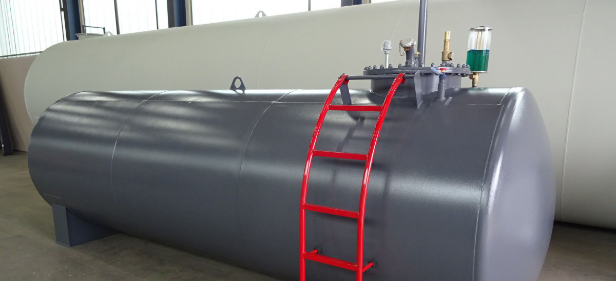 Grauer horizontaler Lagertank mit roten Leiterstufen und Messarmaturen in einer Halle.