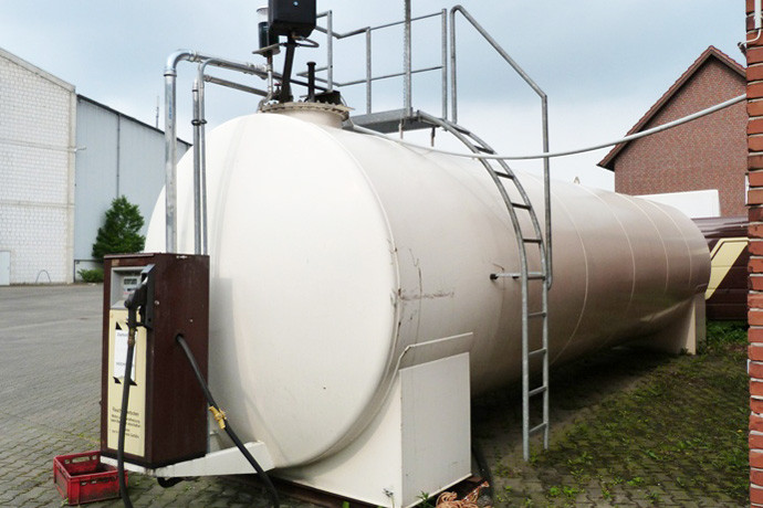 weißer großer zylindrischer Tank mit Leiterpodest und einer Zapsäule steht auf einem Betriebsgelände mit Hallen im Hintergrund