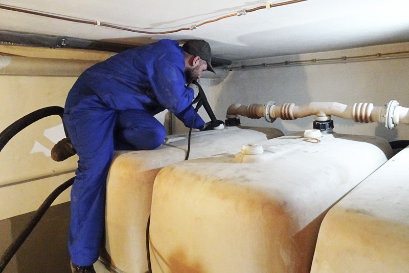 Techniker in blauem Overall arbeitet mit Gerät auf einem Heizöltank im engen Tankraum eines Kellers